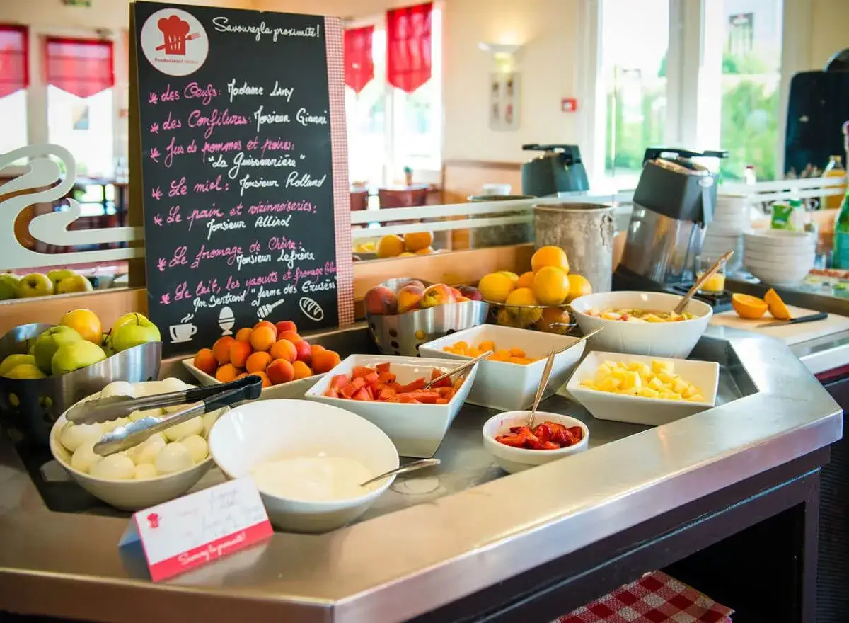 Un buffet de repas du matin avec des fruits frais, des œufs, du fromage et des confitures. Une ardoise liste les produits locaux.