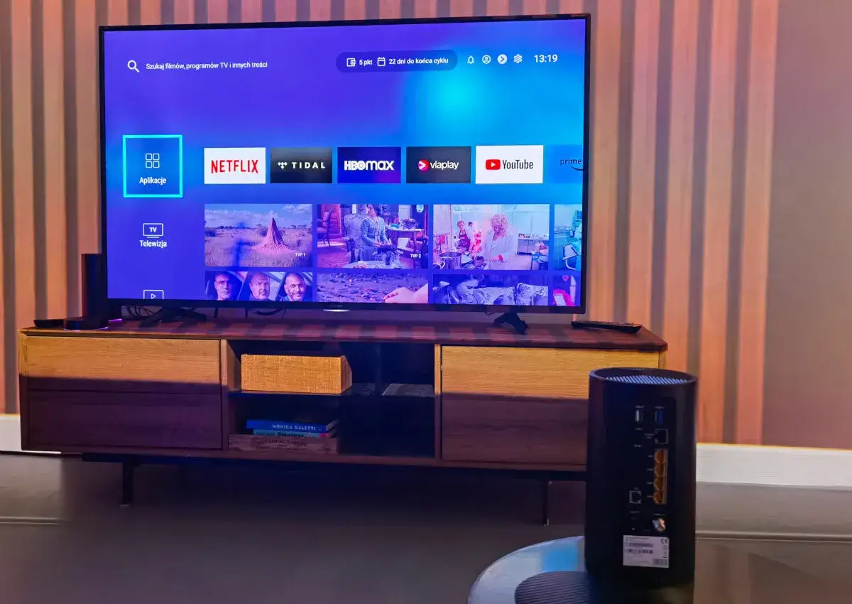 Nowoczesny salon z telewizorem Smart TV, na którym widać aplikacje jak Netflix i YouTube. Obok stoi router, symbolizujący dostęp do internetu od UPC Polska.