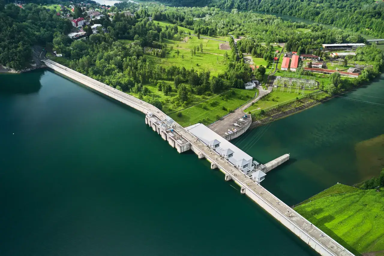 Ogromna zapora na sanie, otoczona zielonymi wzgórzami i lasami, spiętrza turkusową wodę. Na koronie zapory widać ludzi spacerujących.