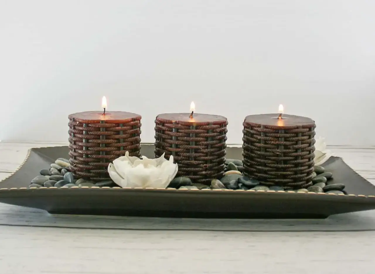 Three lit dripless candles with a woven texture sit on a tray with stones and a white flower.