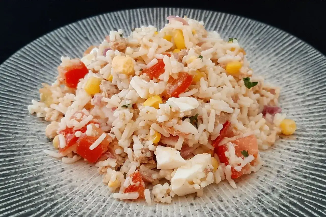 Une généreuse portion de salade de riz thon, garnie de dés de tomates, de maïs et d'œufs durs, sur une assiette texturée.