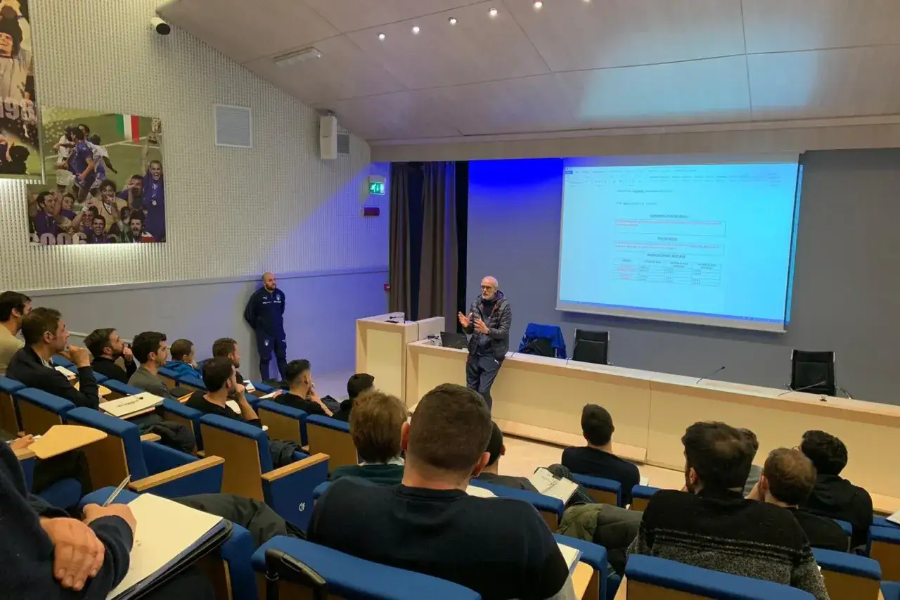 Un relatore spiega dati su un proiettore a un pubblico interessato, un'opportunità per chi vuole lavorare nel calcio.