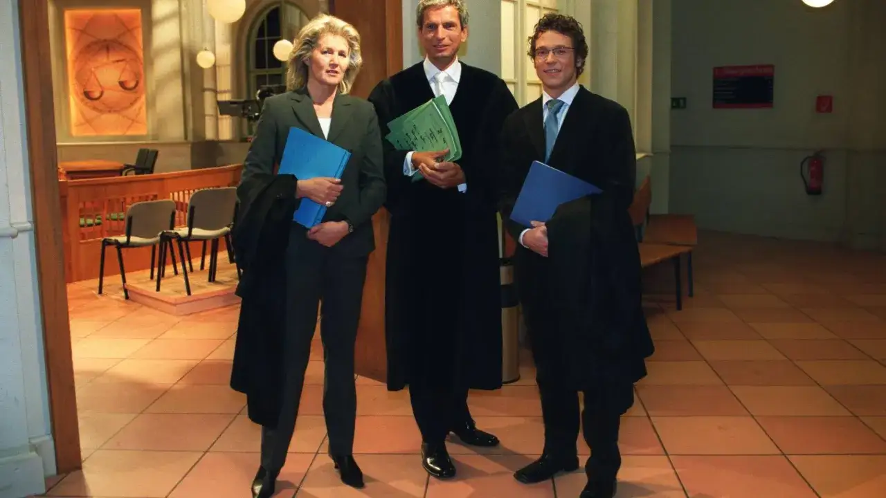 Drei Personen in Roben und Anzügen, die Akten halten, stehen in einem Gerichtssaal. Sie zeigen, wie richtiges Verhalten vor dem familiengericht aussehen kann.