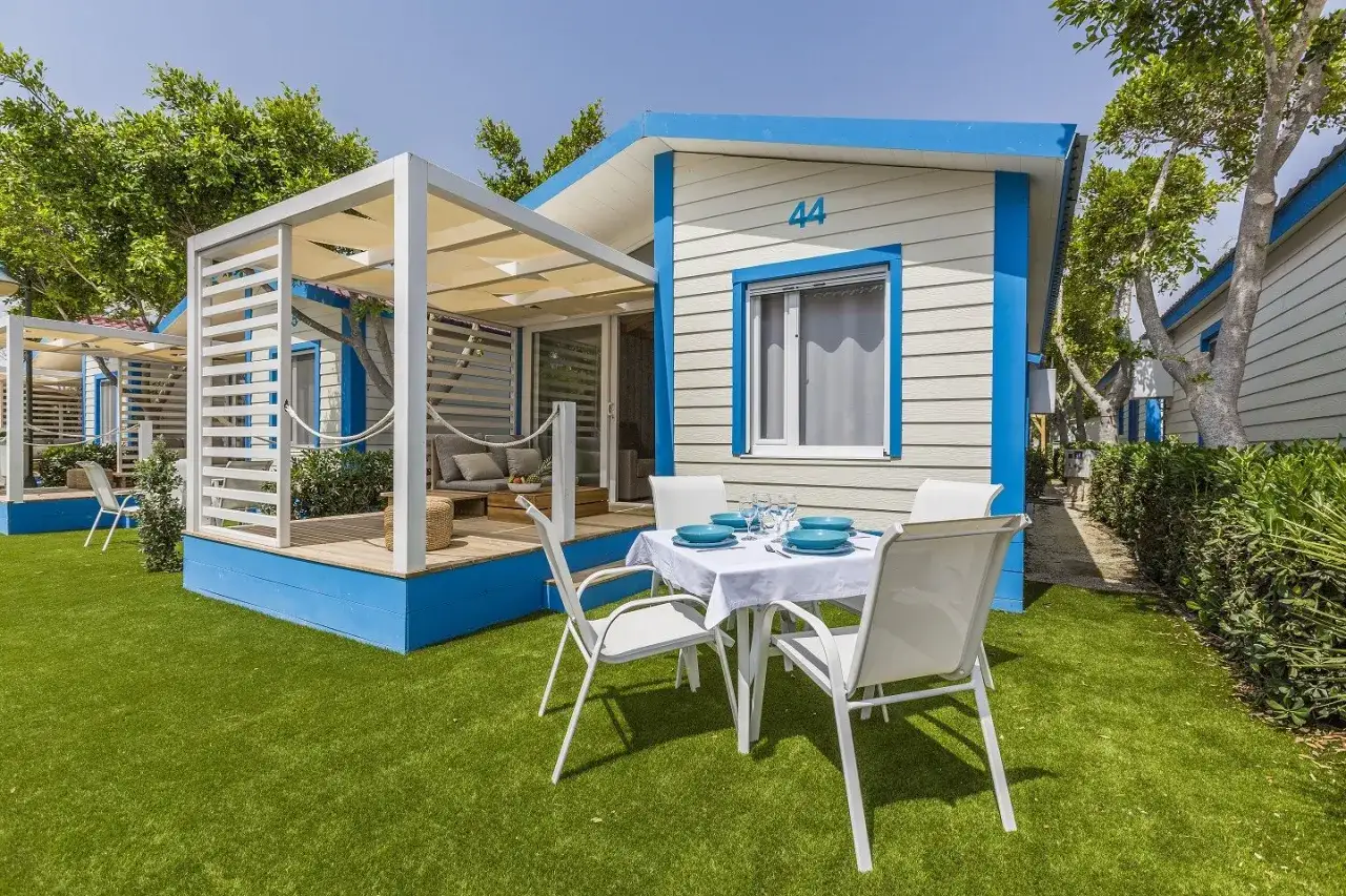 Mesa puesta para cenar al aire libre en el camping Alannia Guardamar. Casas de madera con porche y césped artificial.