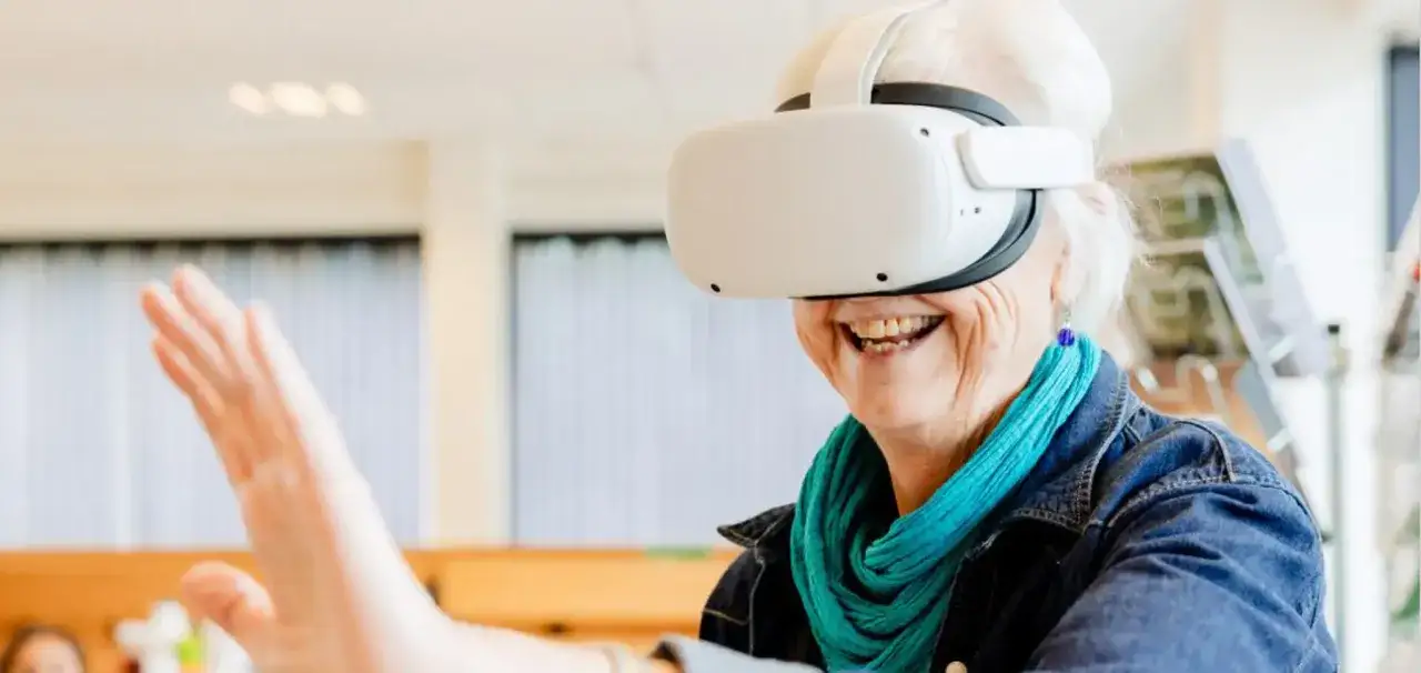 &Auml;ldre kvinna ler med VR-glas&ouml;gon, str&auml;cker ut handen. Ny teknik kan st&auml;rka sociala relationer och minska ensamhet.