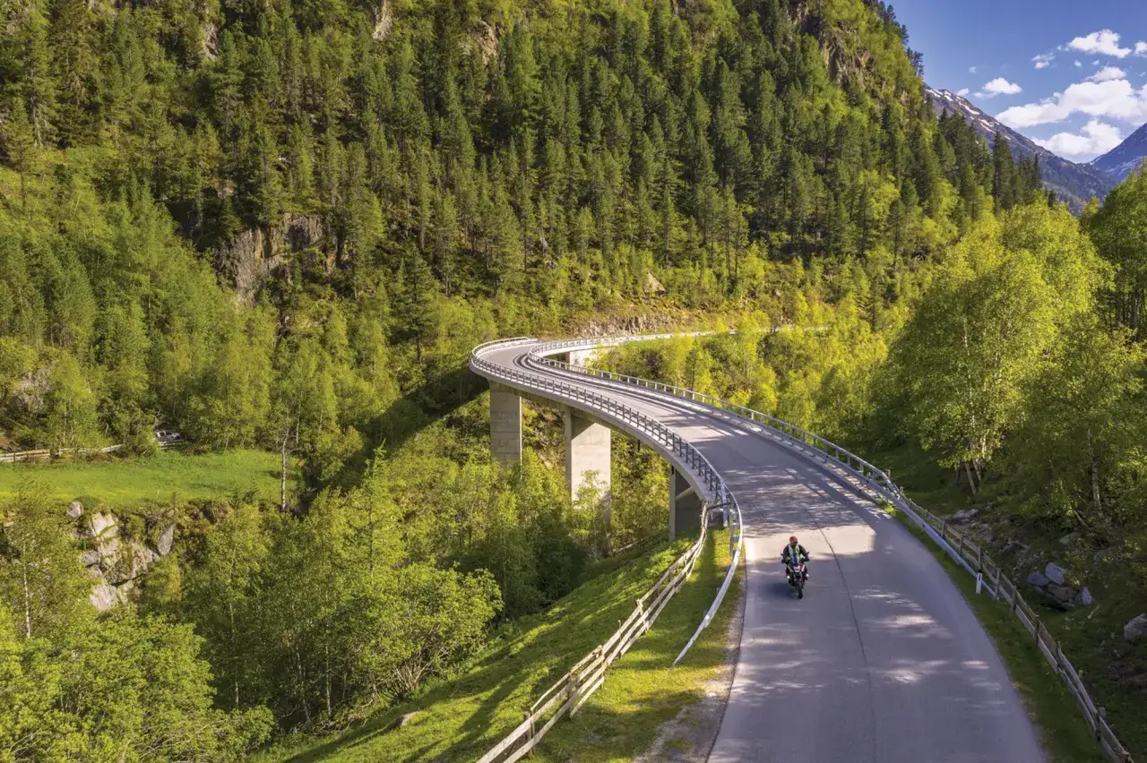 Un motociclista percorre una strada panoramica in Austria, circondato da foreste lussureggianti e montagne.