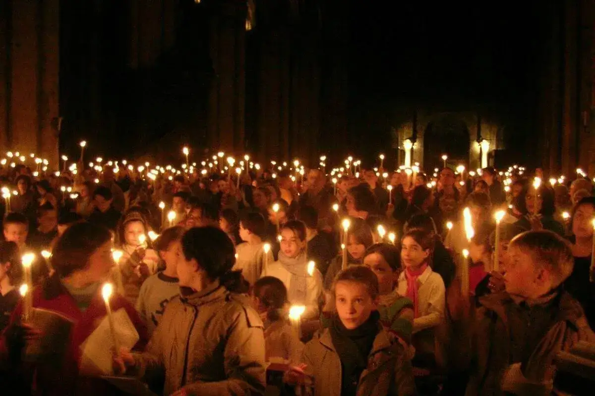 A crowd of people, many children, hold candles aloft in a dark space, participating in a solemn vigil.