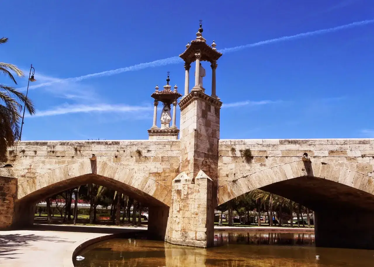 Puente de piedra con estatuas en el **Jardín del Turia**, bajo un cielo azul con estelas de avión.