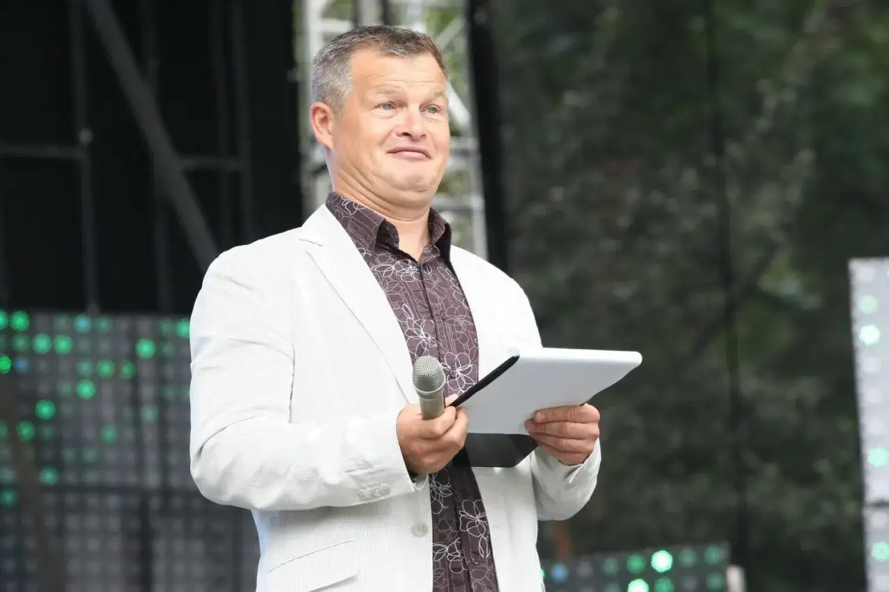 Bieleniinik z TVN, w białej marynarce, trzyma mikrofon i tablet, gotowy do poprowadzenia wydarzenia.