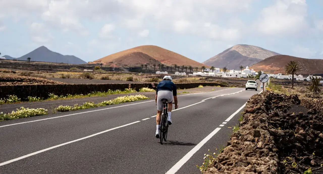 Ein Radfahrer auf einem Rennrad fährt auf Lanzarote durch eine vulkanische Landschaft. Weiße Häuser und Palmen säumen die Straße.