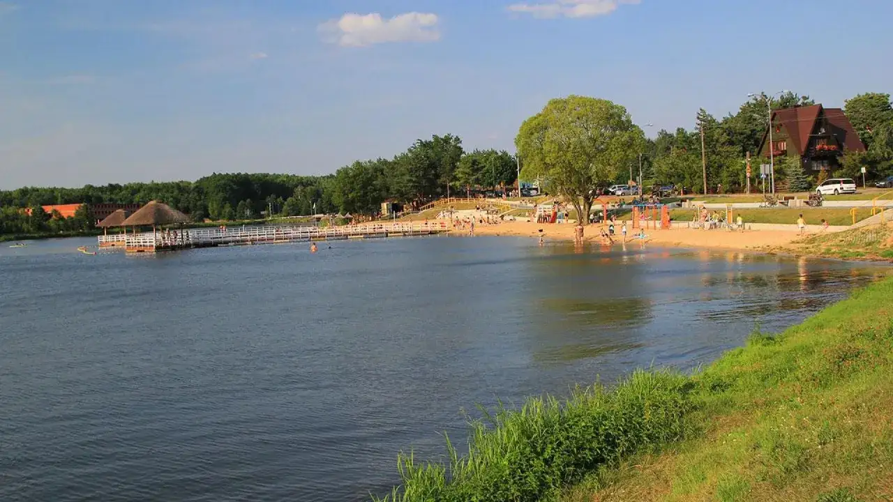 Słoneczny dzień nad jeziorem w Siemiatyczach. Plaża pełna ludzi, pomost z zadaszeniem, drzewa i budynki w tle. Idealne siemiatycze atrakcje.