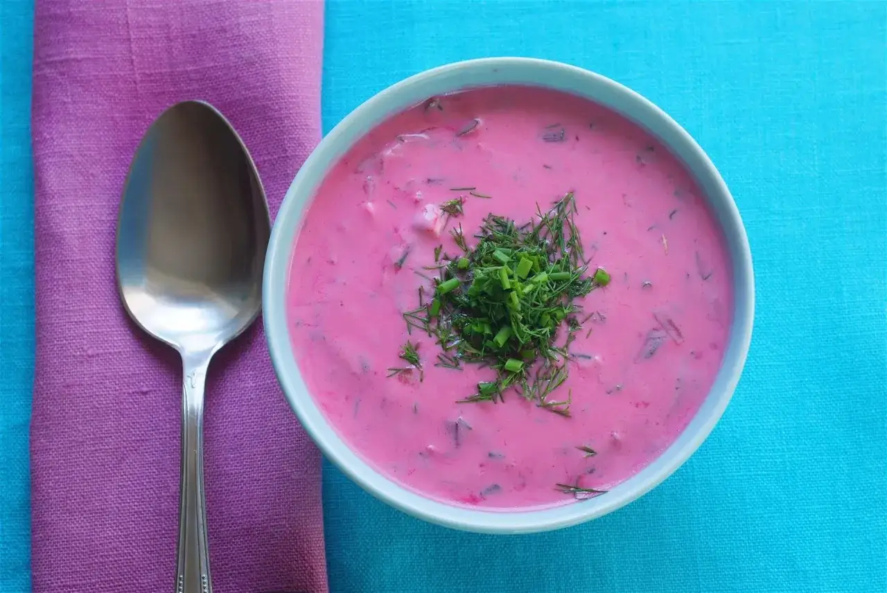 Zuppa fredda di barbabietole, un piatto tipico polacco, guarnita con aneto fresco. Accanto, un cucchiaio e un tovagliolo viola.
