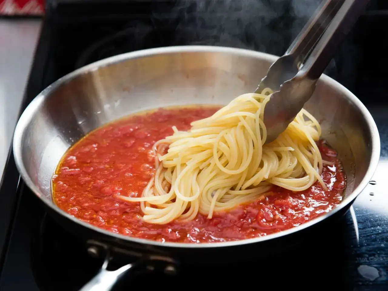 Szczeg&oacute;łowy moment, jak zrobić spaghetti: szczypce dodają ugotowany makaron do gorącego sosu pomidorowego na patelni.