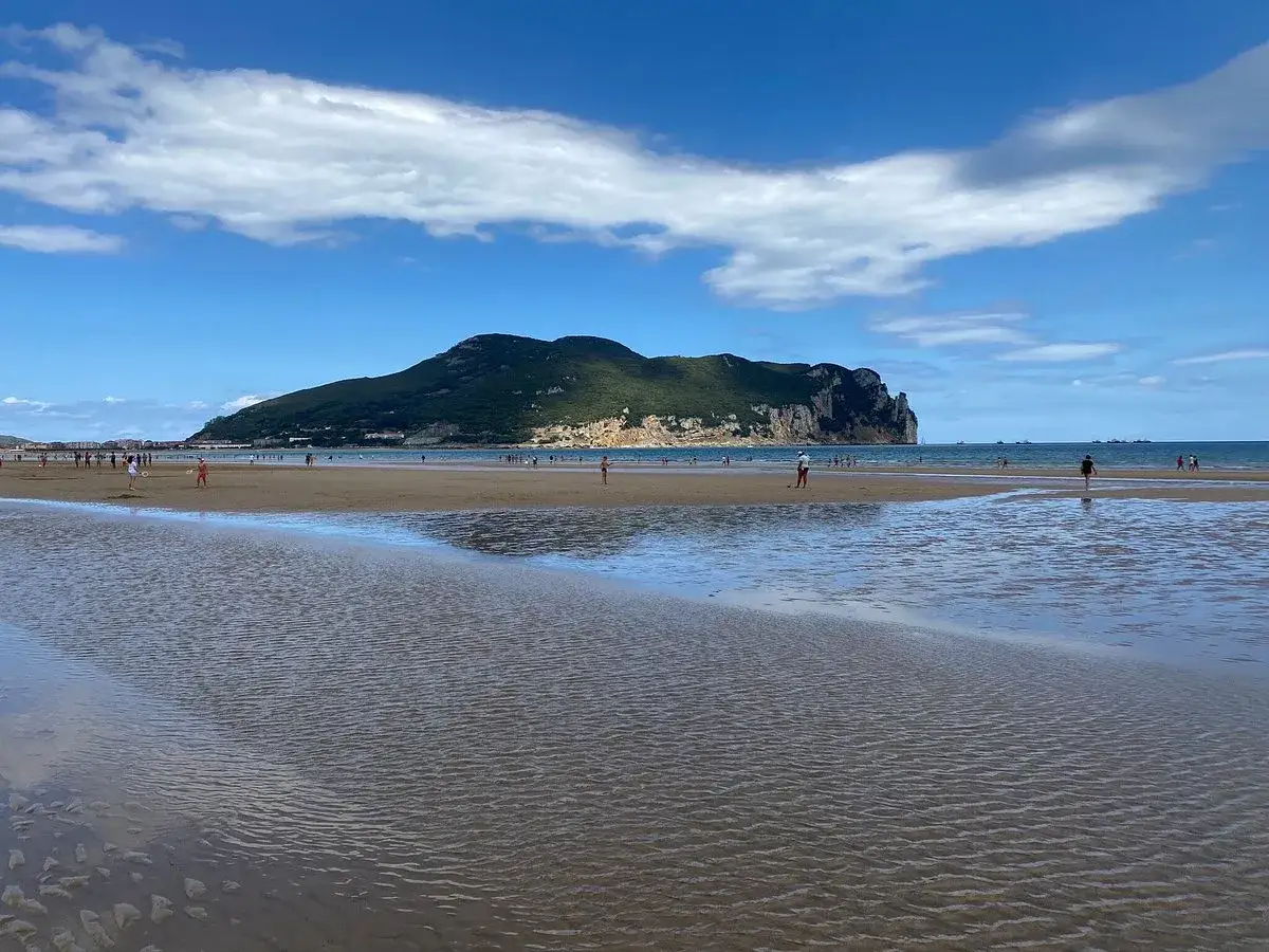 La **playa Laredo** se extiende bajo un cielo azul con nubes blancas. Personas pasean por la arena mojada, mientras una gran montaña verde se alza al fondo.