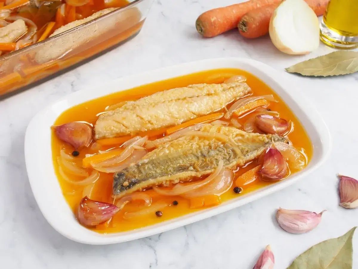 Dos filetes de caballa en escabeche, con zanahoria, cebolla y ajos, en una fuente blanca.