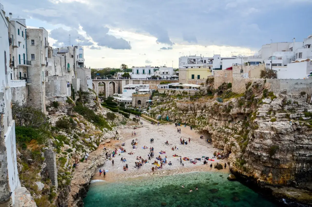Malownicze Polignano a Mare z białymi domami na klifach, kamienistą plażą pełną ludzi i turkusową wodą.
