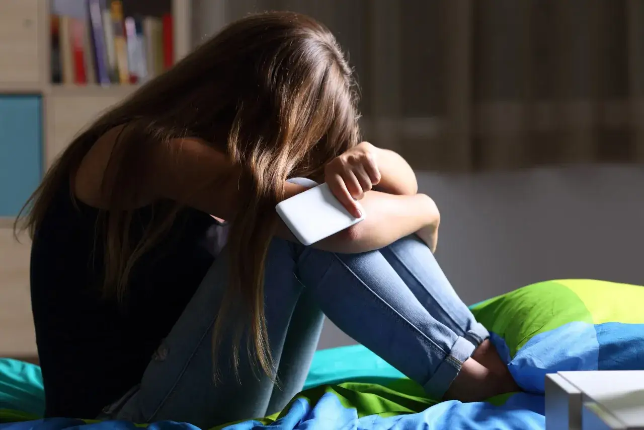 Dziewczyna siedzi skulona na ł&oacute;żku, trzymając telefon. Wok&oacute;ł niej wszystko się układa, tylko nie jej.