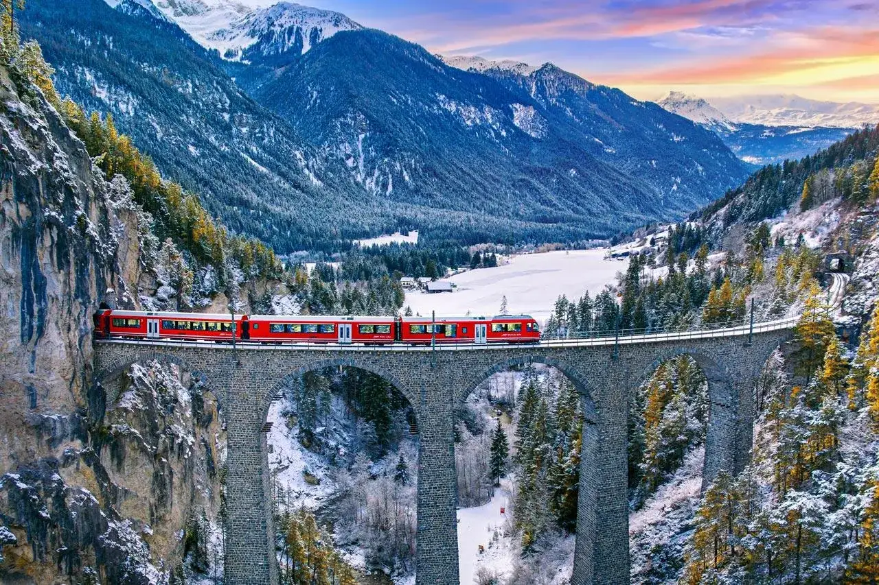 A red train crosses a stone viaduct in a snowy Swiss valley, on its way from Zermatt to Andermatt.