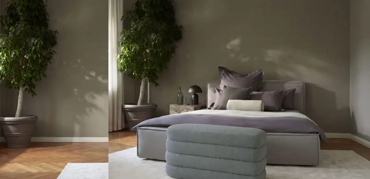 Ein modernes Schlafzimmer in gedeckten Farben mit zwei gro&szlig;en Pflanzen, einem Bett mit vielen Kissen und einem Hocker.