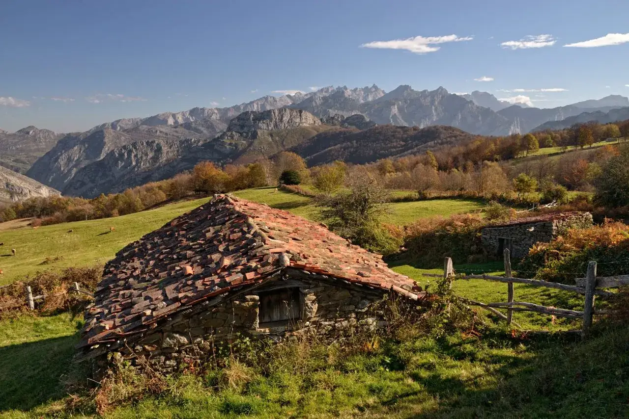 Majestuosas montañas en el **parque natural de Ponga**. Un antiguo refugio de piedra con tejado de tejas rojas se alza en un prado verde, con otras construcciones al fondo.