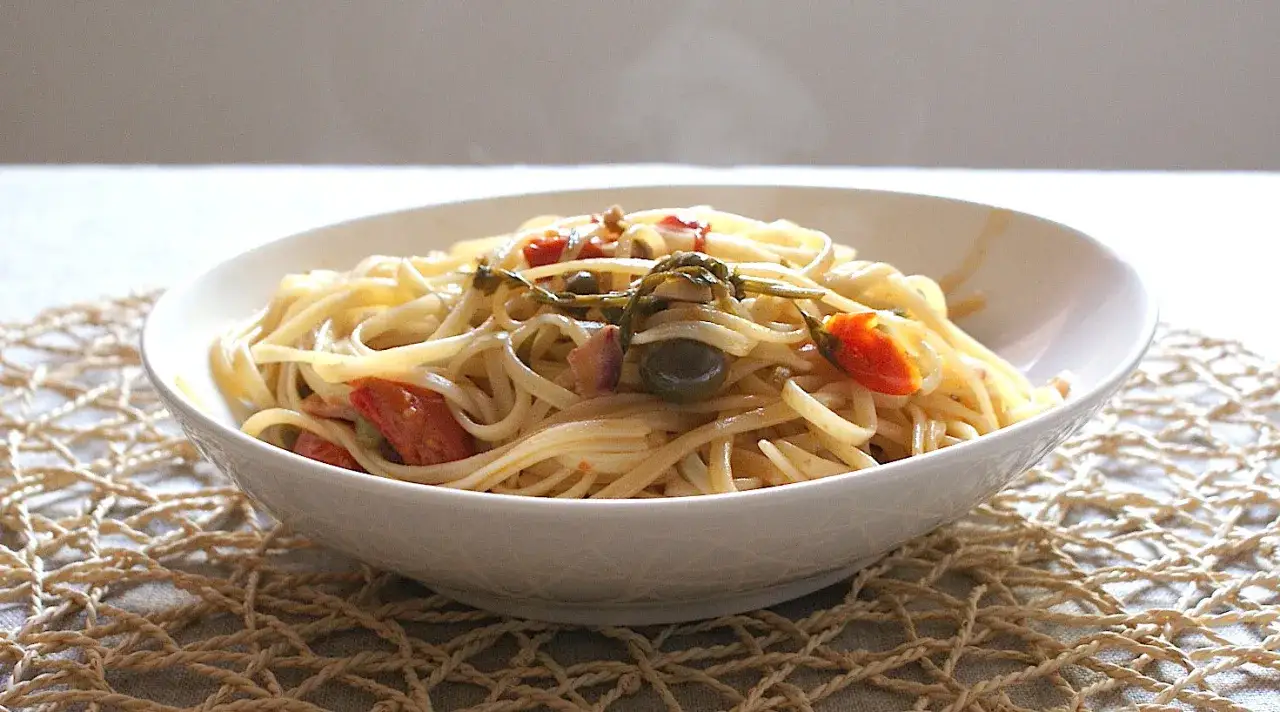 Un plato de pasta con calamares, tomates y aceitunas, servido en un cuenco blanco sobre un mantel tejido.