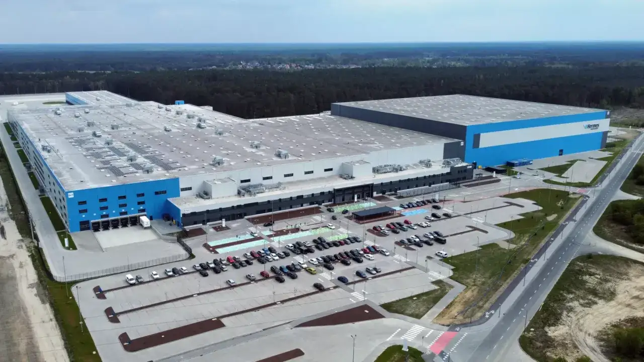 Ogromny magazyn Hermes w Niemczech, z niebieskimi elementami i logo firmy. Obok parking pełen samochodów i droga.