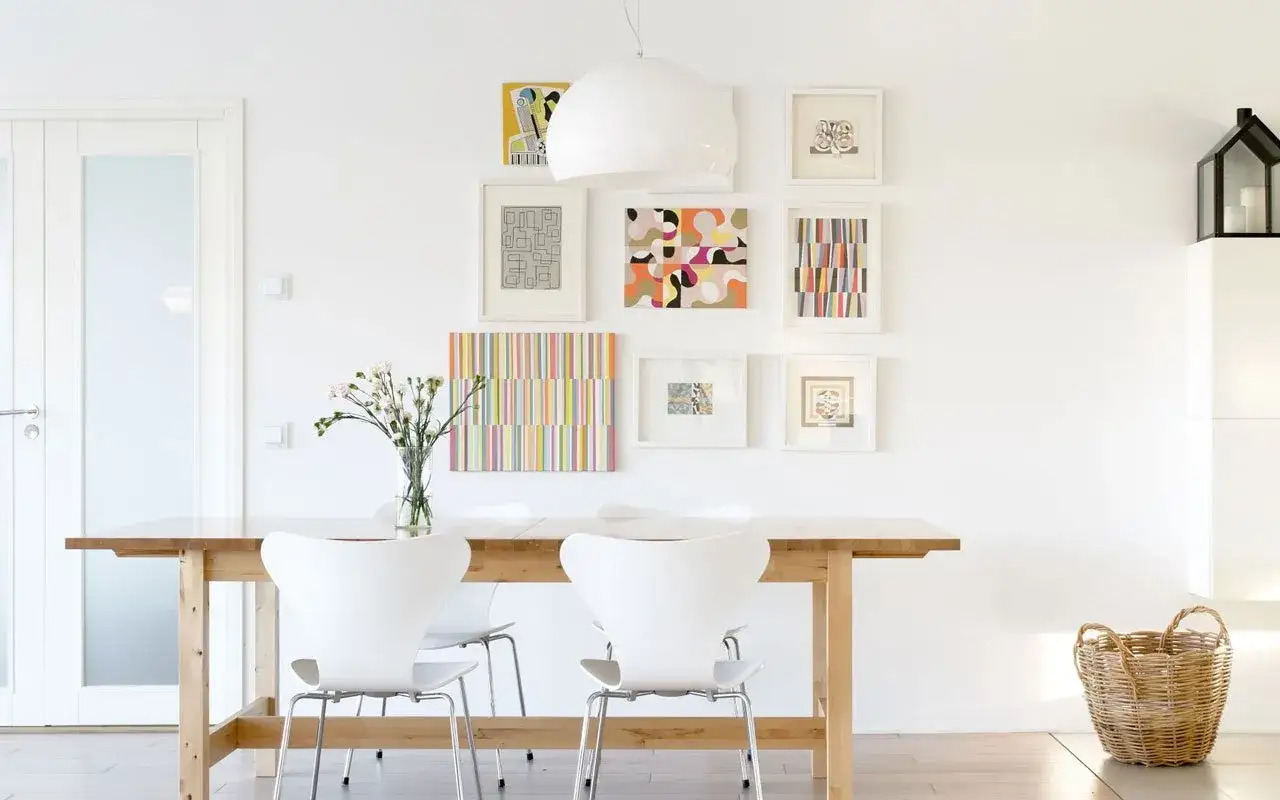 Galleria di quadri colorati per come riempire una parete vuota in cucina. Tavolo in legno e sedie bianche completano l'arredo.