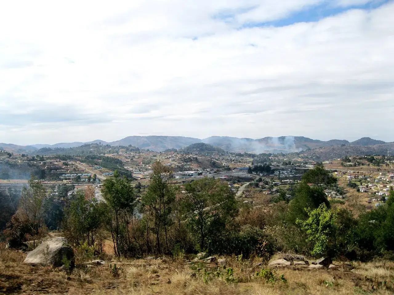 Krajobraz z Mbabane, Eswatini. Wzgórza pokryte zabudowaniami i zielenią, z dymem unoszącym się w oddali.