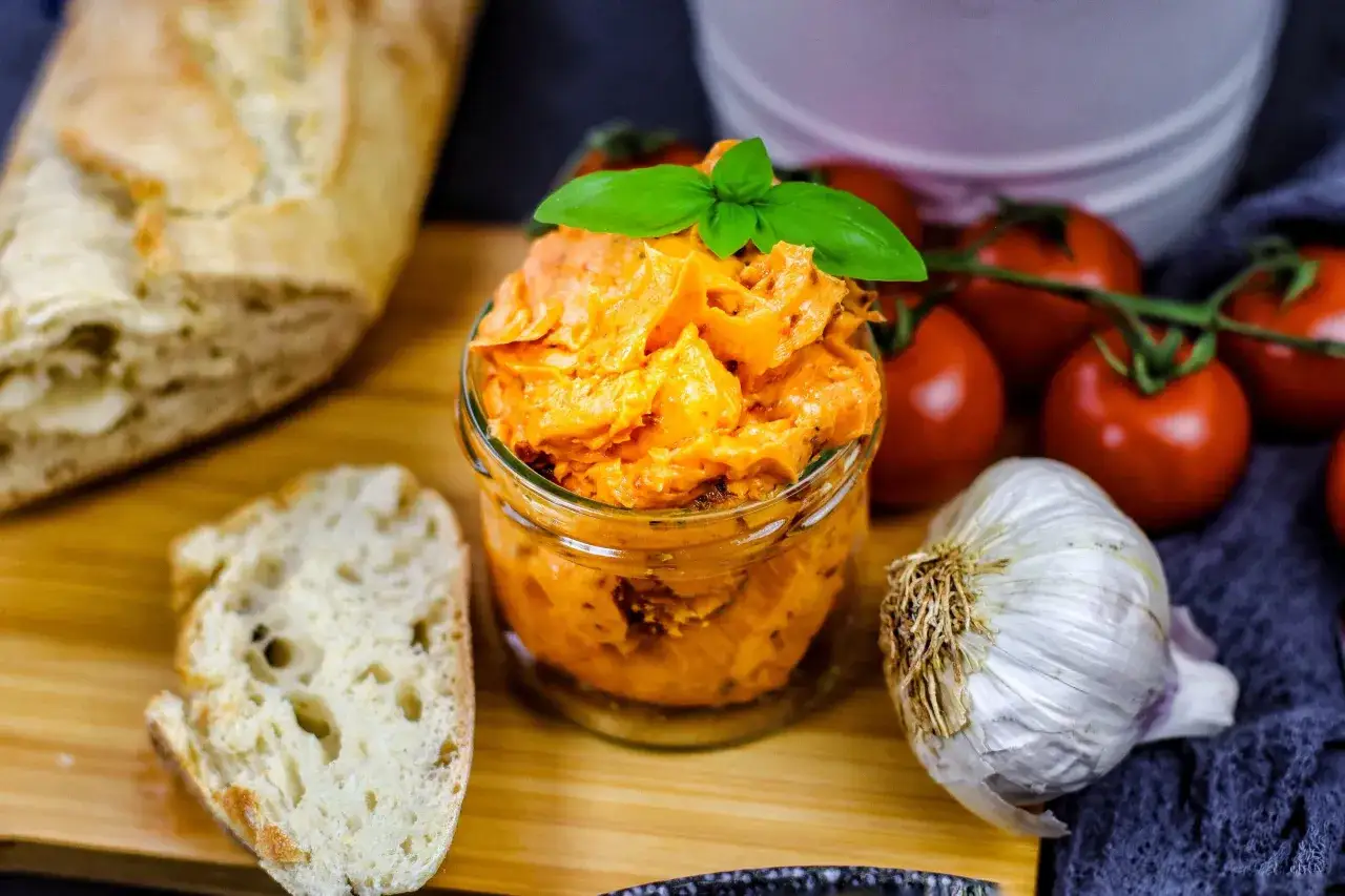 Glas mit Tomatenbutter, garniert mit Basilikum. Daneben Baguette, Knoblauch und frische Tomaten. Perfekt für ein Tomatenbutter Rezept.