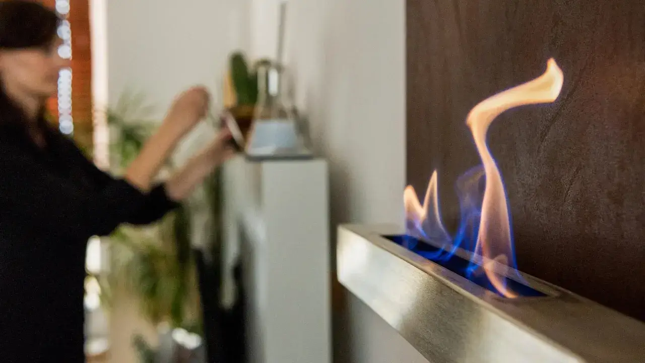 Eine Person befüllt einen Ethanol-Kamin, bei dem die Ethanol gefahren beachtet werden müssen. Die Flammen tanzen in Blau und Orange.