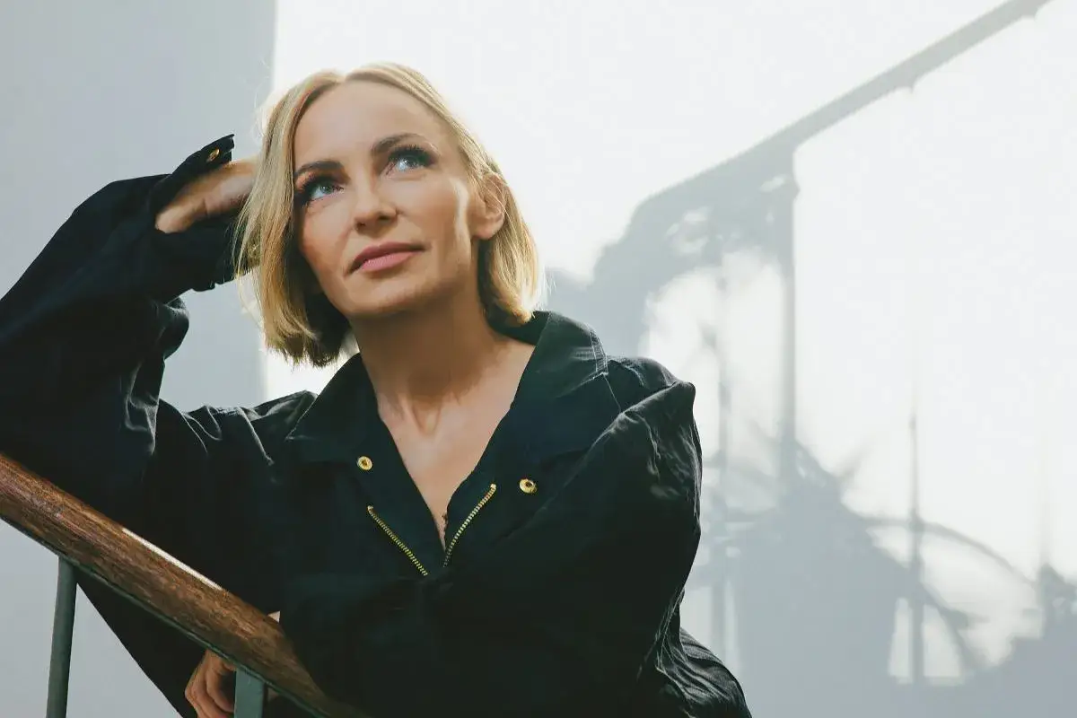 Ania Wyszkoni, w wieku, w którym doświadczenie maluje się na twarzy, opiera się o poręcz schodów, patrząc w górę z zadumą.