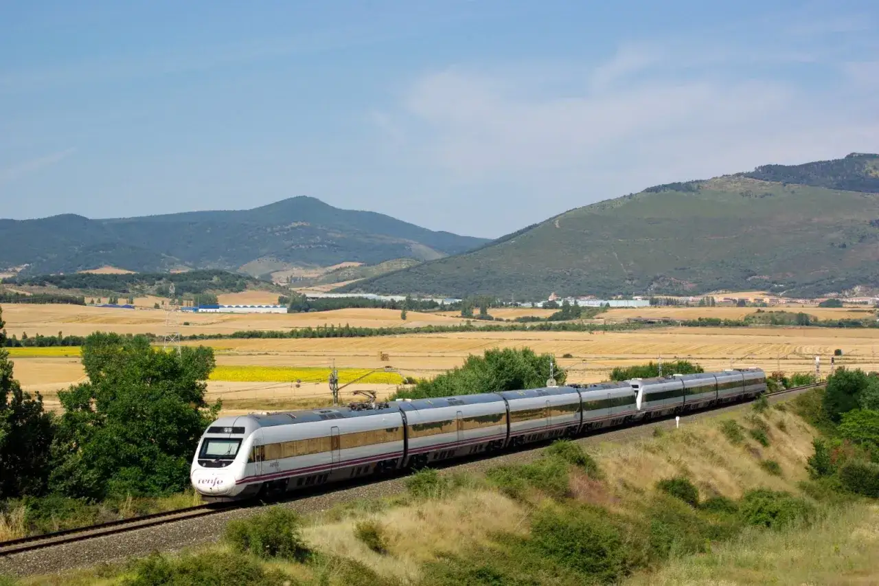 Un moderno tren de alta velocidad Renfe recorre un paisaje rural con campos dorados y monta&ntilde;as verdes.