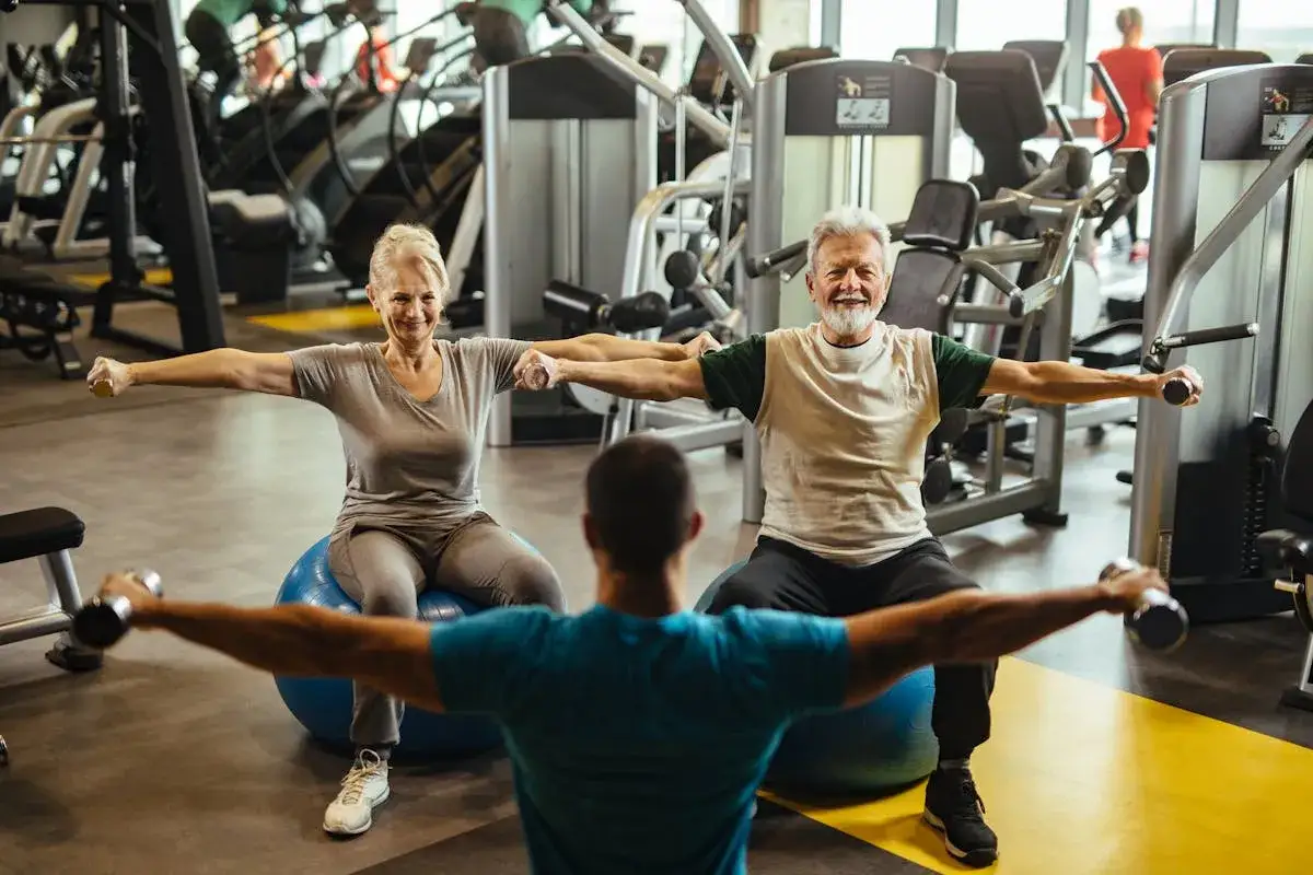 Un couple de retraités fait un exercice ballon gym avec des haltères, guidé par un coach.