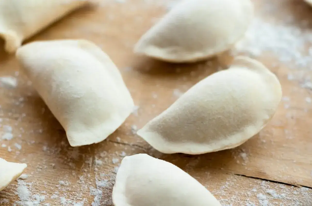 Surowe pierogi leżą na drewnianej desce, posypane mąką. To idealne ciasto na pierogi bez jajka, gotowe do nadziewania.