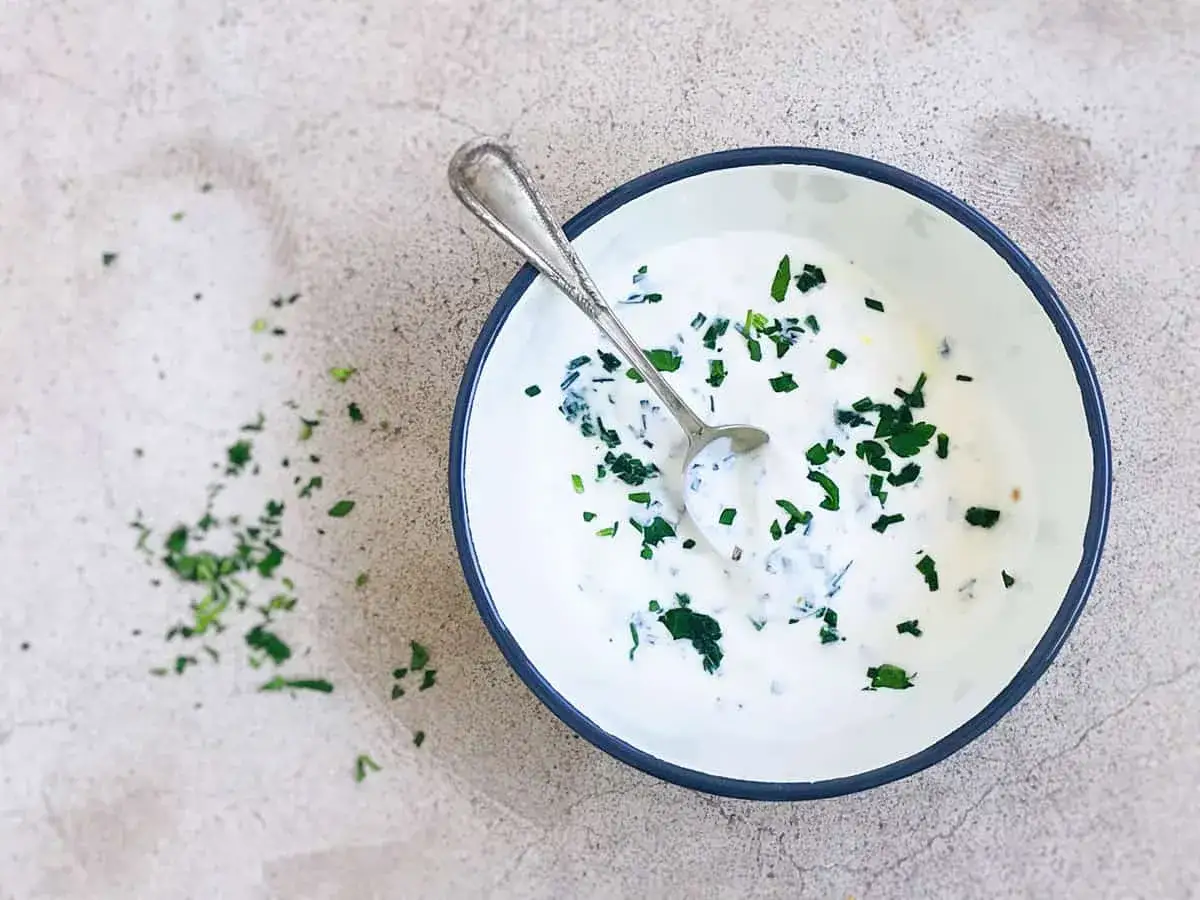 Un cuenco con salsa de yogur vegana y hierbas frescas picadas, con una cuchara metálica.