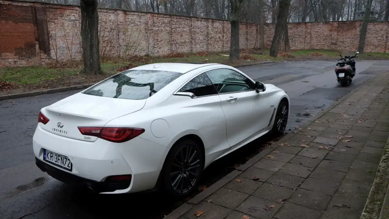 Białe Infiniti Q60s stoi na mokrej ulicy obok ceglanego muru.