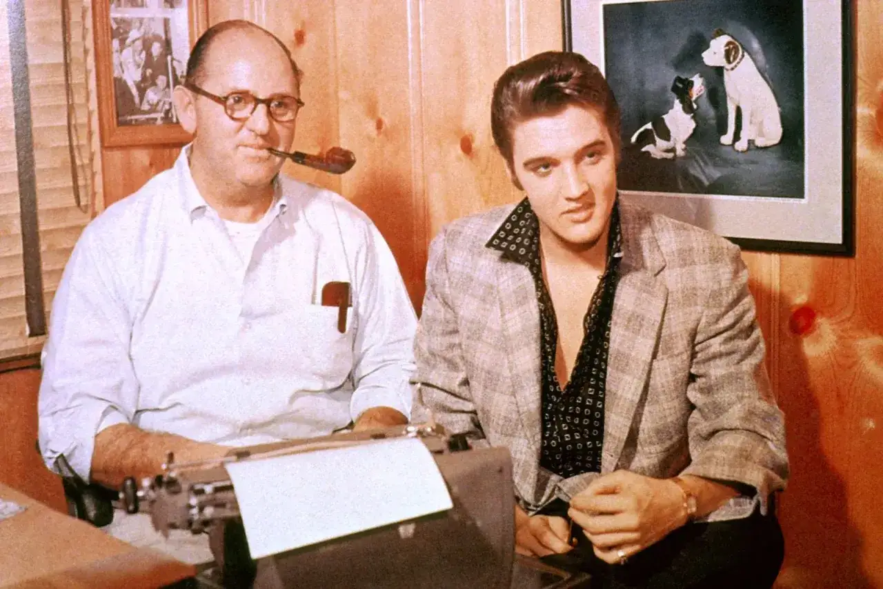Elvis Presley und Colonel Parker, sein Manager, sitzen nebeneinander. Parker raucht Pfeife, Elvis blickt nachdenklich.