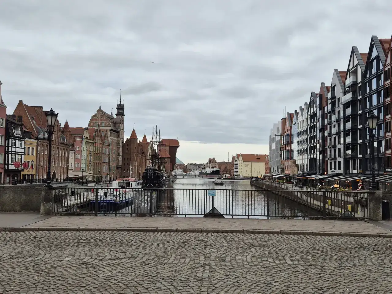 Widok na Motławę z kamiennego bruku, z kolorowymi kamienicami po lewej i nowoczesnymi budynkami po prawej. W oddali widać żaglowiec i statki.