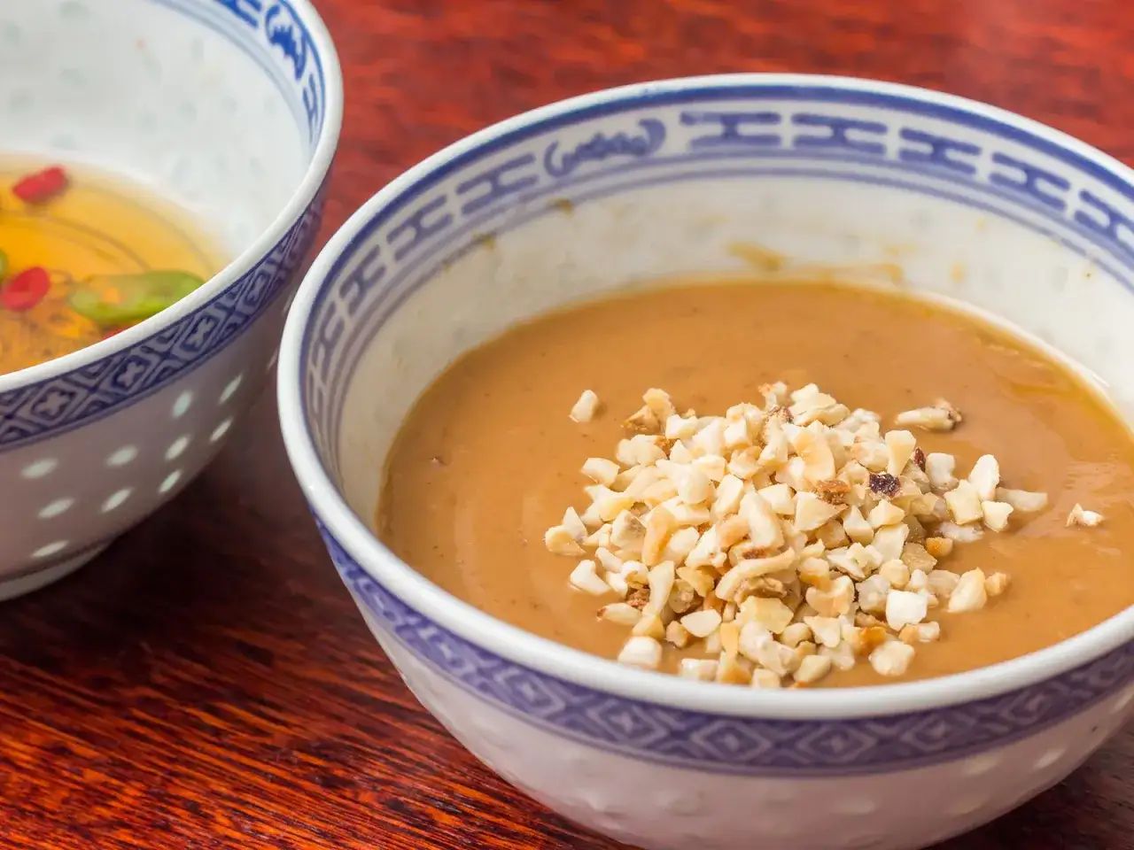 Cremige Erdnuss Sauce selber machen, garniert mit gehackten Erdnüssen in einer Schüssel mit blau-weißem Muster.