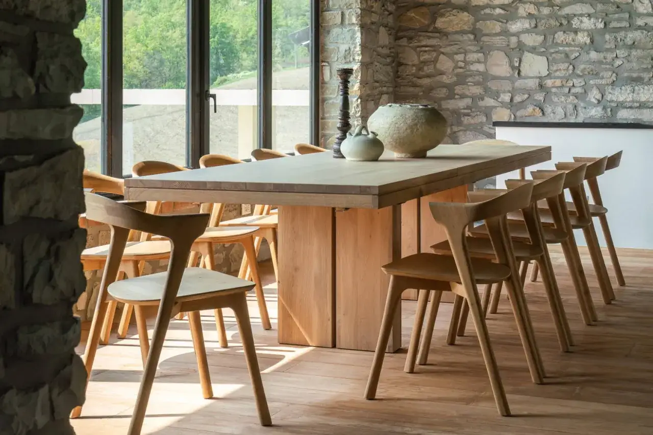 A spacious, light wood dining table for 12, surrounded by matching chairs, is set against a rustic stone wall and large windows.