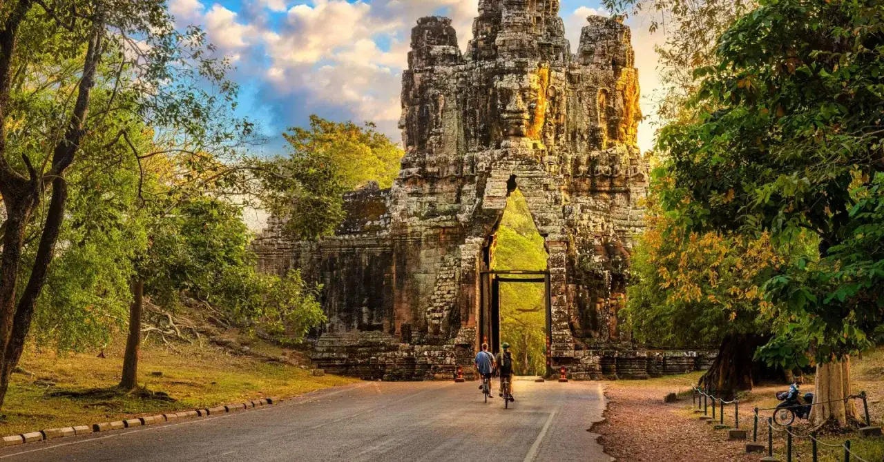 Two cyclists approach a monumental stone gate, one of the ancient Cambodia temples, bathed in golden sunlight.