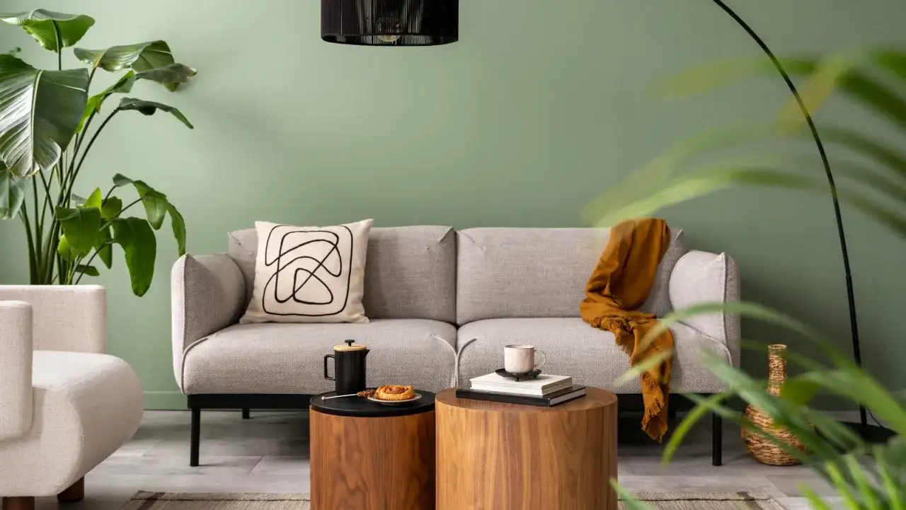 Sof&aacute; gris con coj&iacute;n abstracto y manta naranja, rodeado de plantas y mesas de madera. Tonos fr&iacute;os predominan en la decoraci&oacute;n.