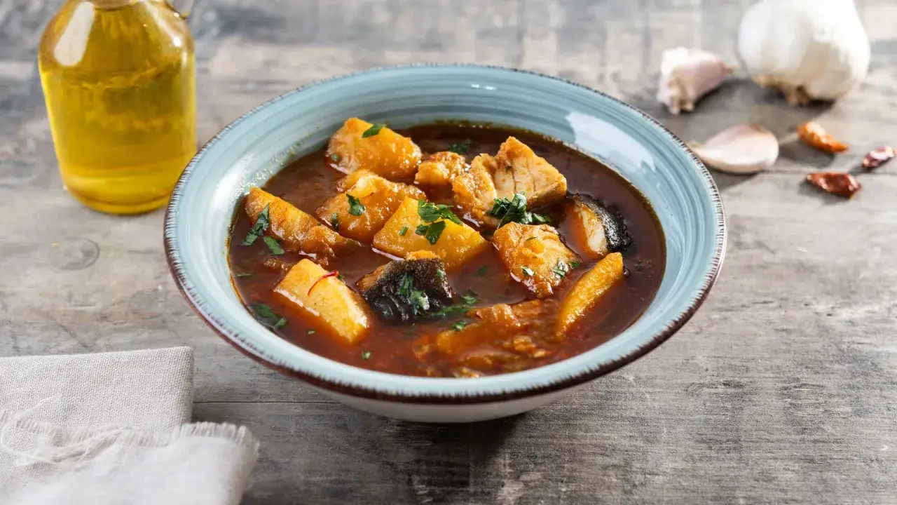 Un tazón de patatas con bacalao en salsa, con aceite de oliva y ajos de fondo.