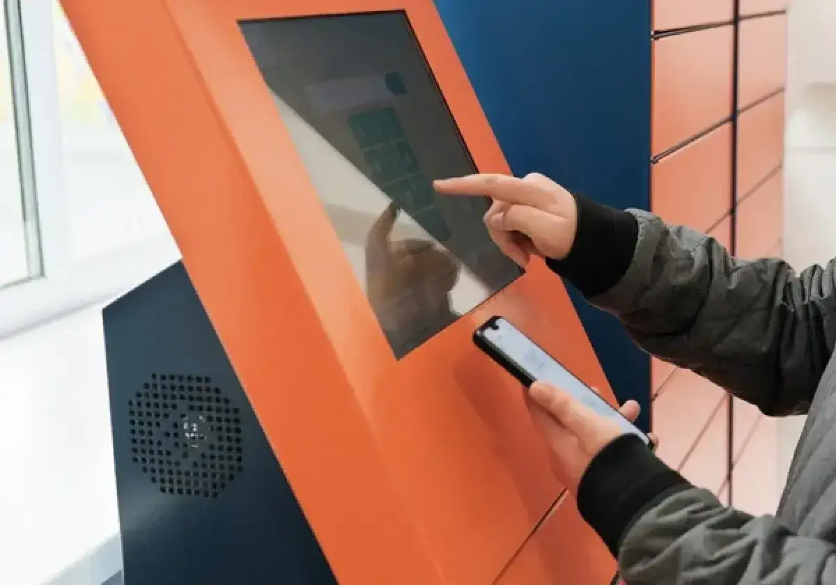 Osoba korzysta z ekranu dotykowego paczkomatu InPost, trzymając telefon. Nie trzeba drukować etykiety, wystarczy kod z aplikacji.