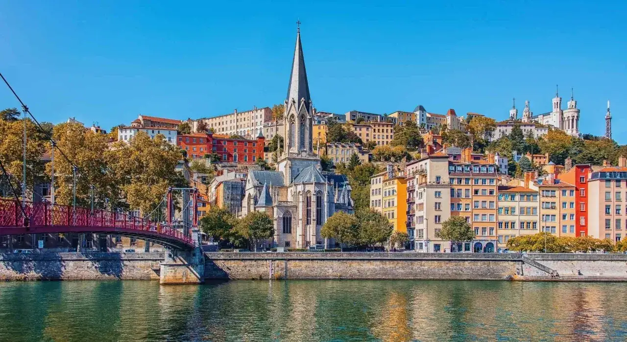 A vibrant cityscape of Lyon, France, with colorful buildings cascading down a hill towards a river. A gothic church with a tall spire dominates the center, a red bridge on the left. Perfect for Lyon travel inspiration.