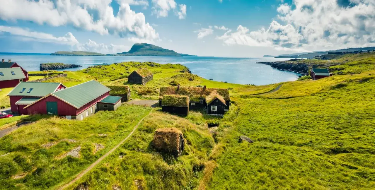 Case tradizionali con tetti d'erba e case rosse, tipiche degli abitanti delle Isole Faroe, immerse nel verde con il mare sullo sfondo.