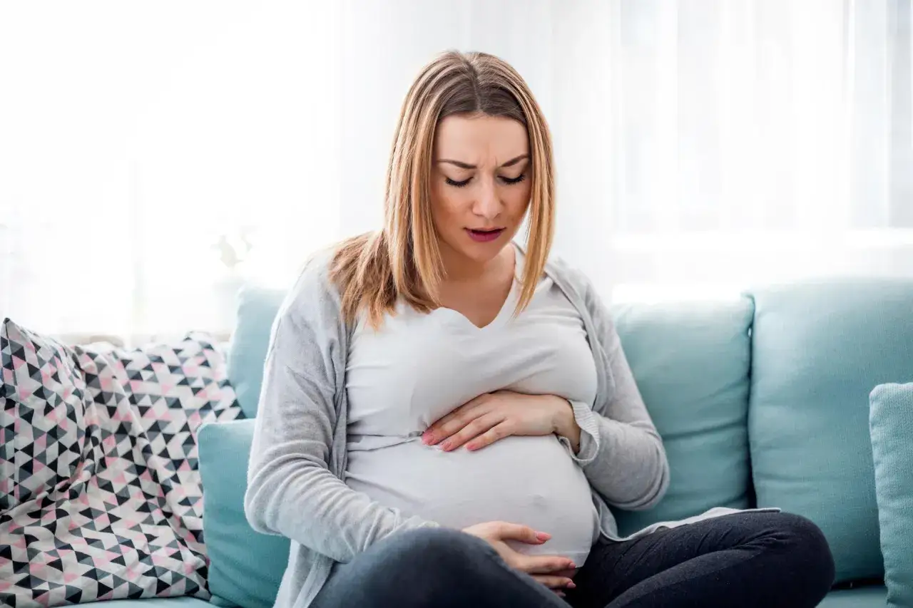 Ciężarna kobieta siedzi na kanapie, trzymając się za brzuch. Jej twarz wyraża dyskomfort, prawdopodobnie czuje skurcze przepowiadające.