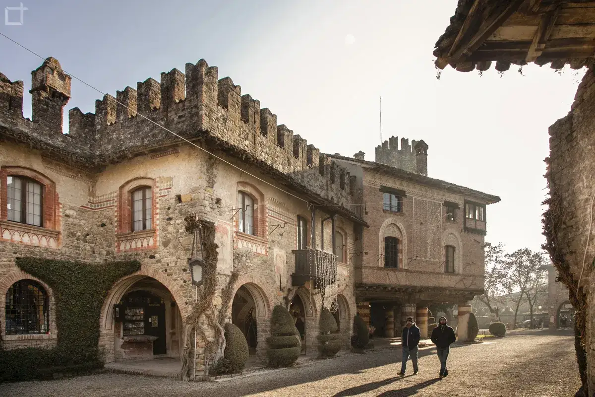 Antico borgo Visconti a Piacenza, con torri merlate e facciate in pietra. Due uomini passeggiano nel cortile illuminato dal sole.