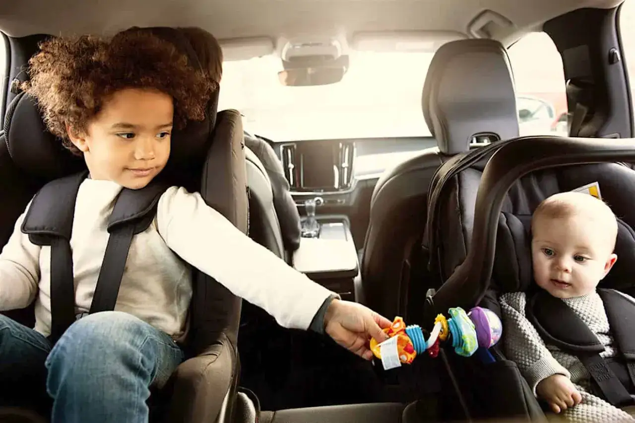 Dos niños, uno mayor y un bebé, viajan seguros en sus sillas de coche. La mayor le pasa un sonajero al bebé, ambos niños mayores a contramarcha.
