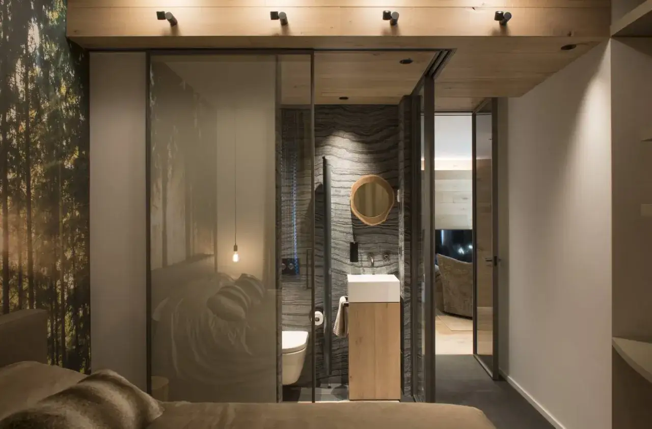 Un moderno bagno in camera da letto con pareti in pietra, lavabo in legno e un letto con vista su una foresta.